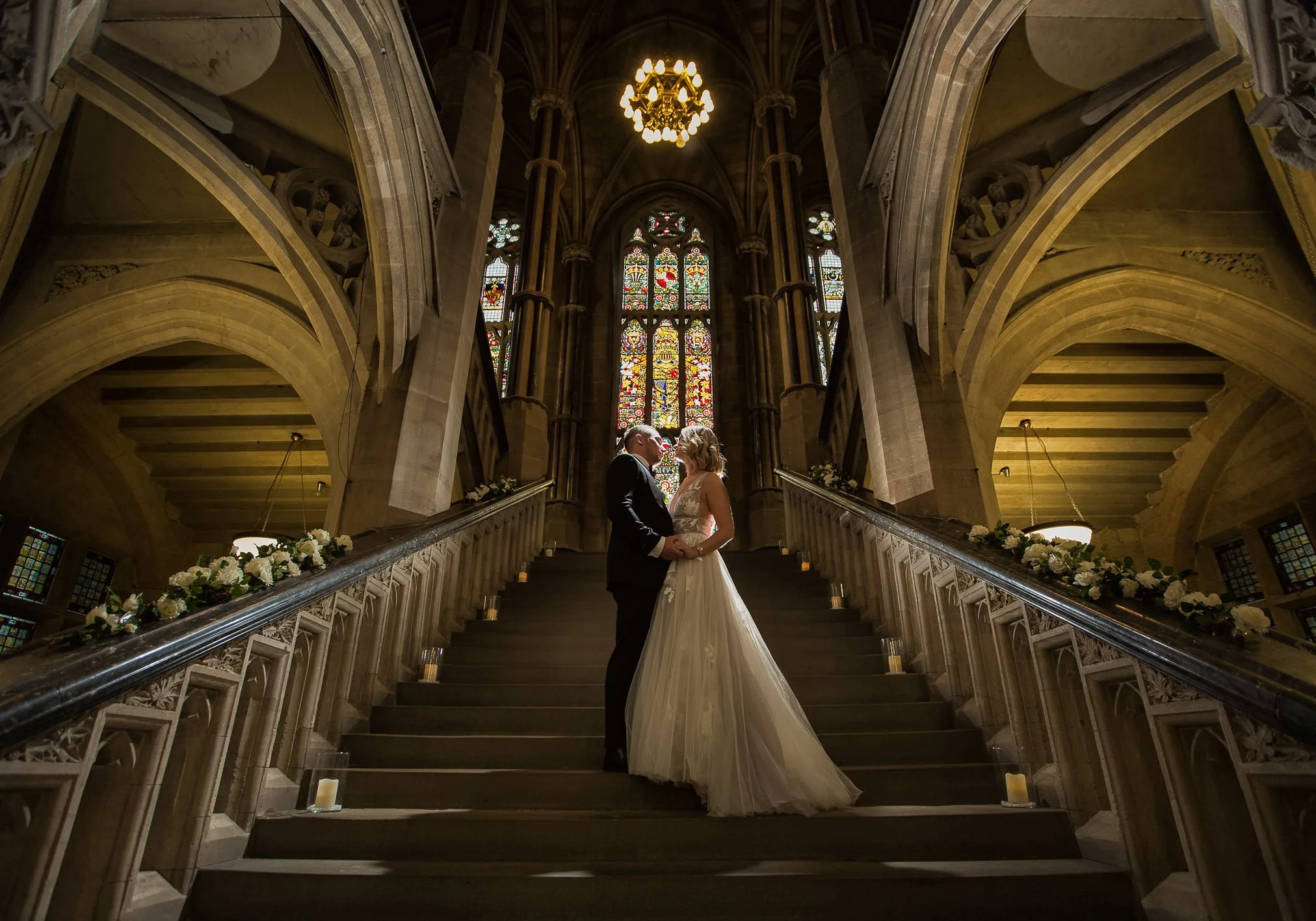 Dynamic scene of bride and groom standing and embracing each other on a stone stepped staircase within a huge grand ornate town hall.