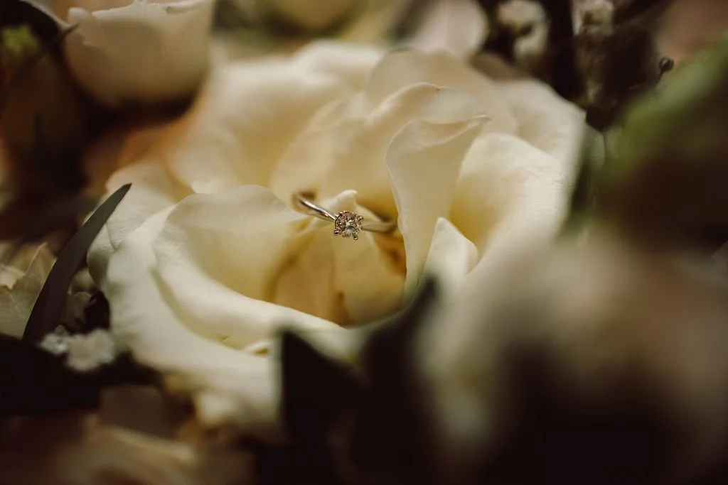 A diamond engagement ring placed inside a white rose surrounded by other flowers.