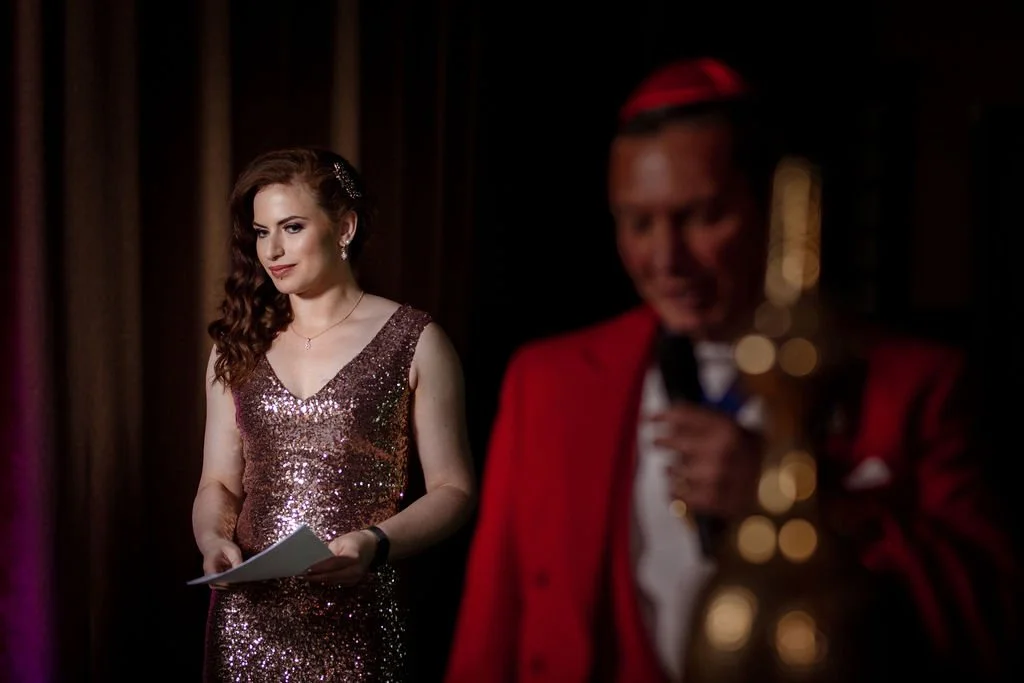 A woman in a sparkly dress holding a paper, standing on stage, with a man in a red jacket and cap, out of focus, holding a microphone in the background.