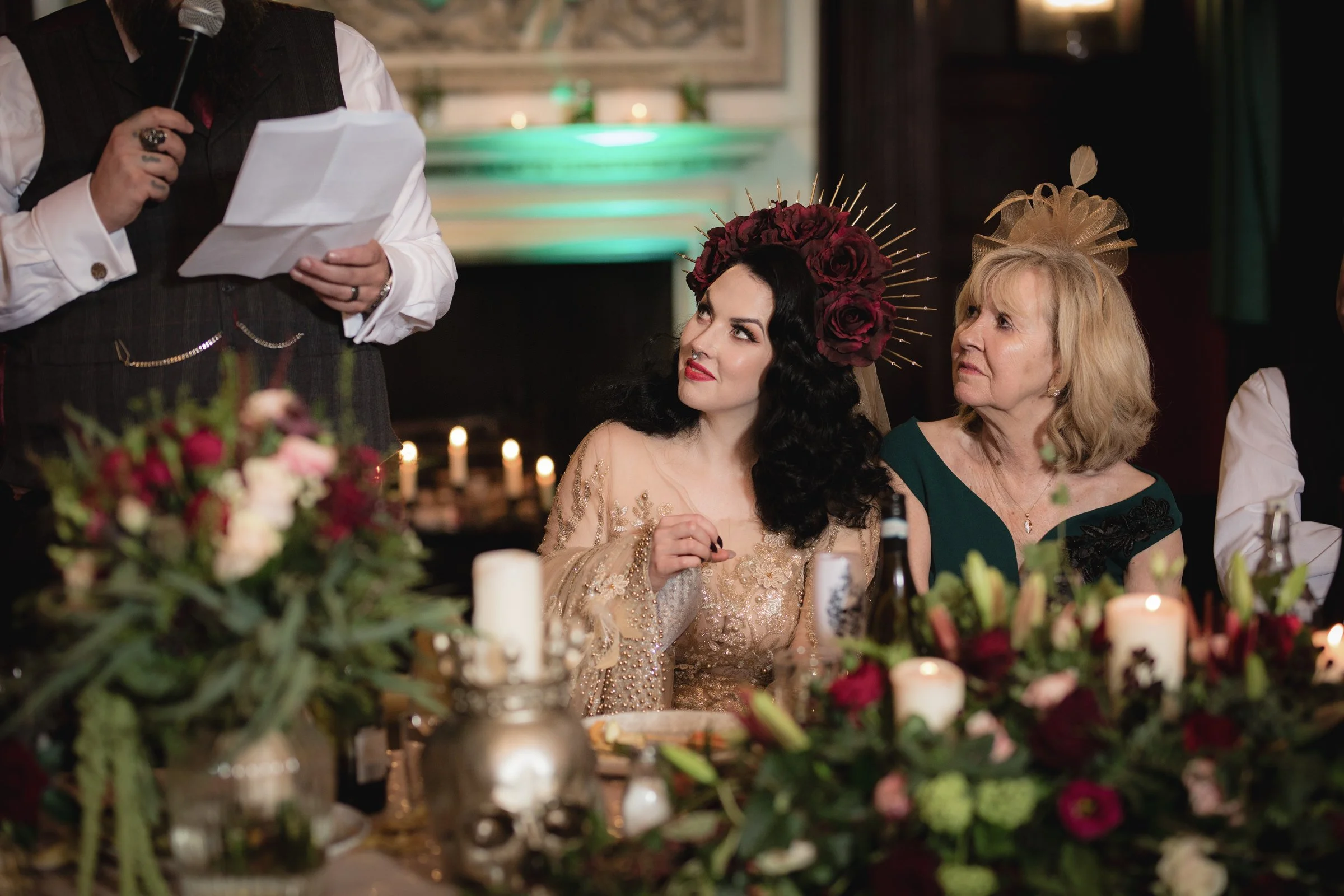 Women attending a formal event, sitting at a decorated table with candles and floral arrangements, listening to a person speaking.