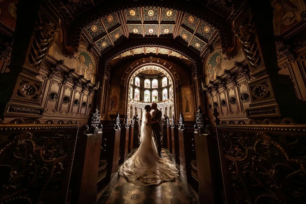 A bride and groom standing in the aisle of a richly decorated church, silhouetted against stained glass windows.