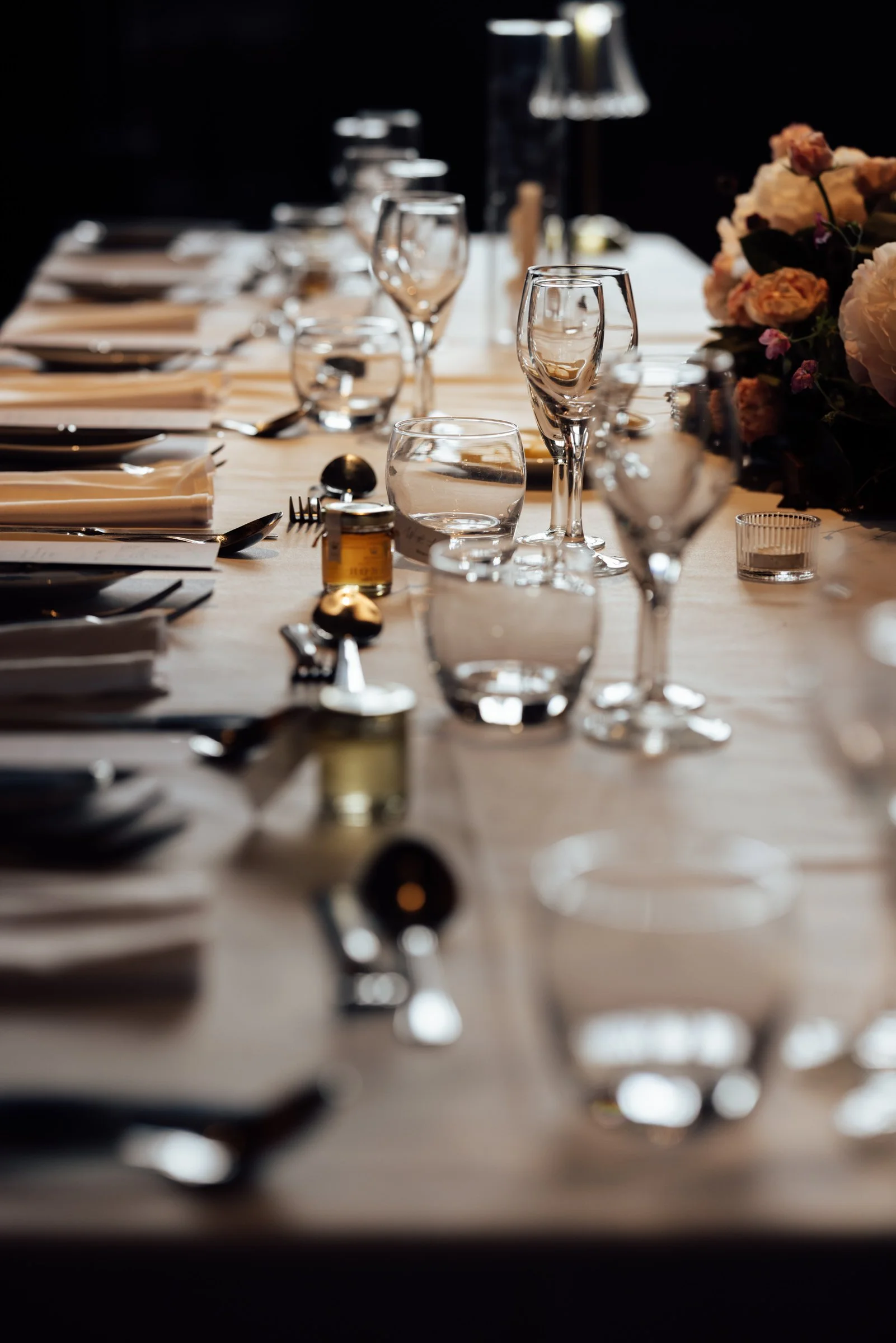 A dining table set for a formal meal with wine glasses, water glasses, plates, silverware, a floral centerpiece, and a small bottle of oil or vinegar.