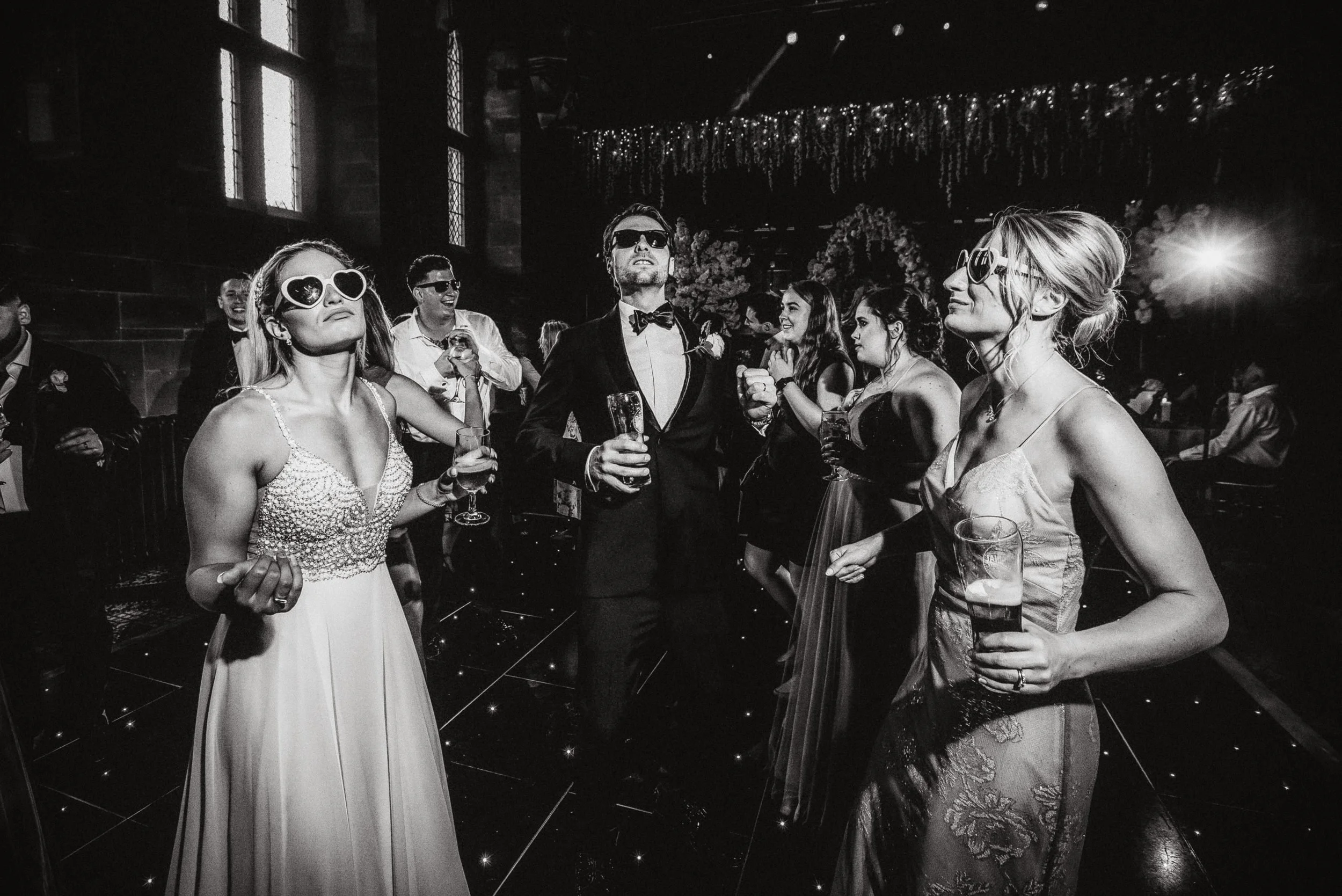 People dancing and celebrating at a wedding reception, wearing sunglasses, with some holding drinks, in a dimly lit decorated venue.