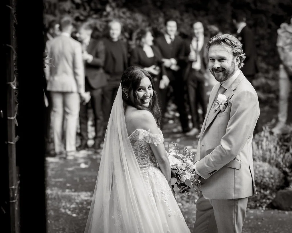 A bride and groom smiling at each other during their wedding ceremony outdoors, with wedding guests in the background.