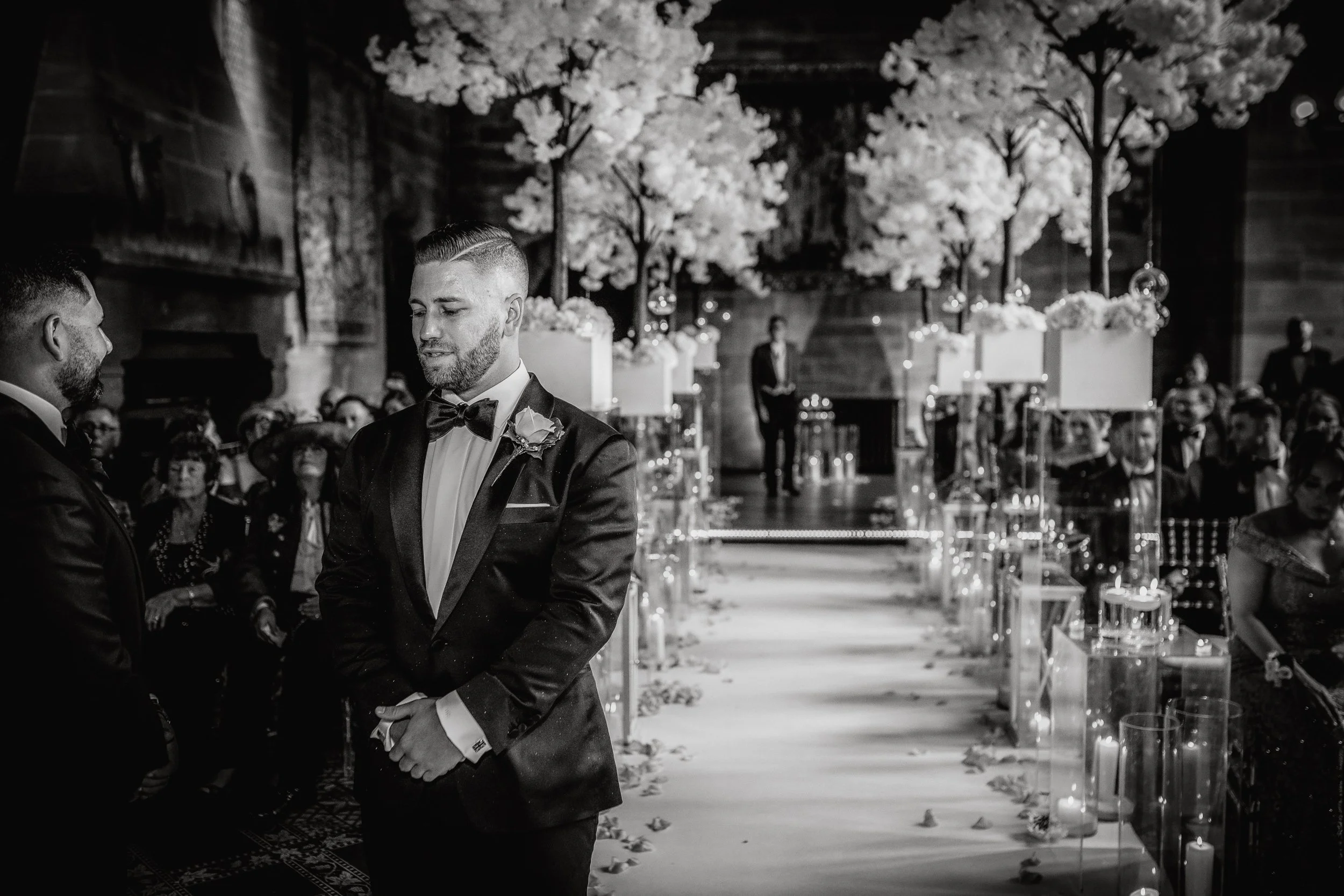 A black-and-white photo of a wedding ceremony. The groom, dressed in a tuxedo with a bow tie, is standing in front of the altar with his eyes closed. Guests sit on either side of the aisle, which is decorated with flower petals, floral arrangements, 
