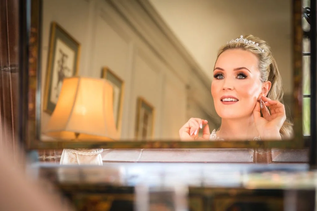 A woman wearing a tiara and earrings, smiling while looking at herself in a mirror.