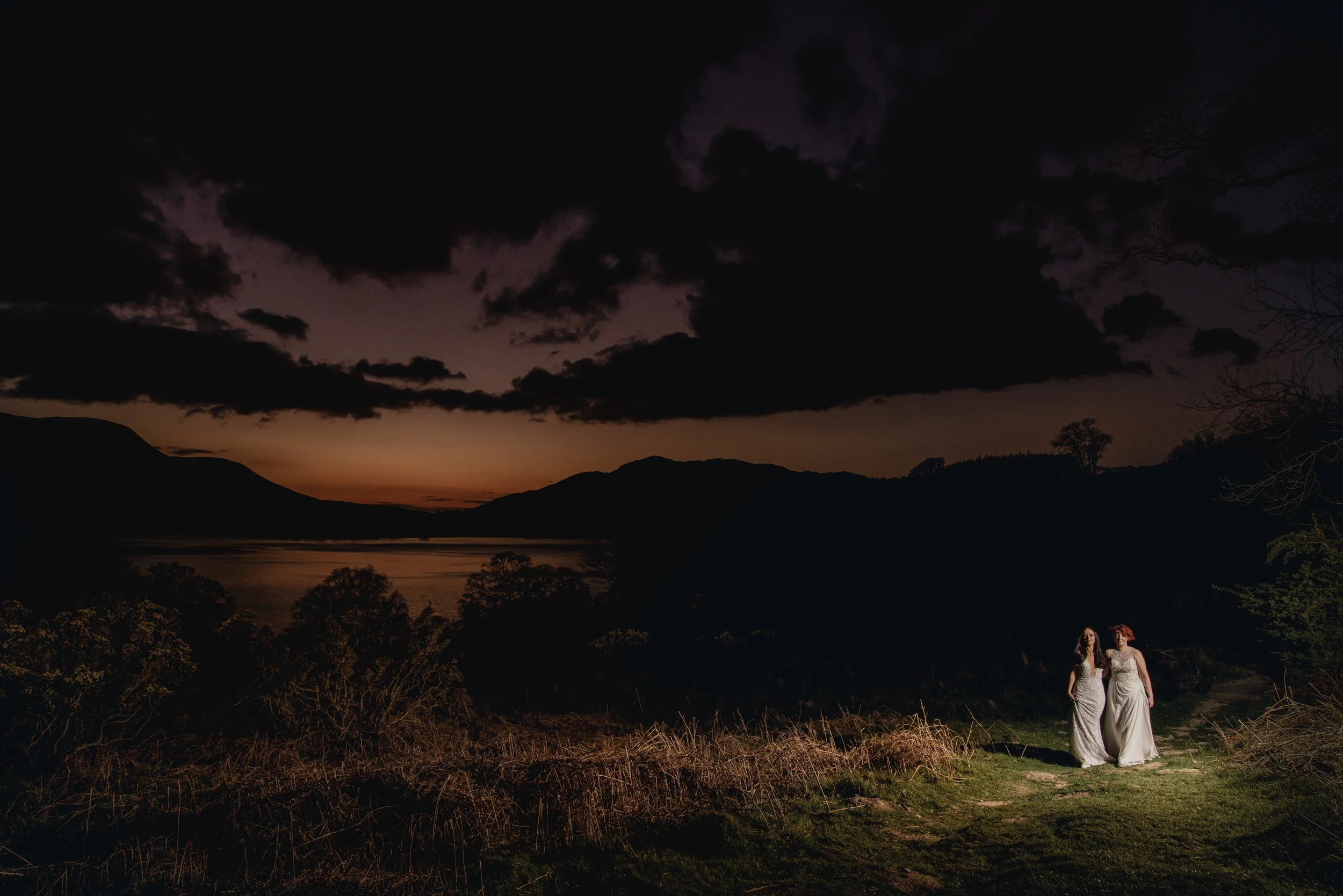 Two women dressed in white gowns walking through a dark natural landscape during sunset with mountains and trees in the background.