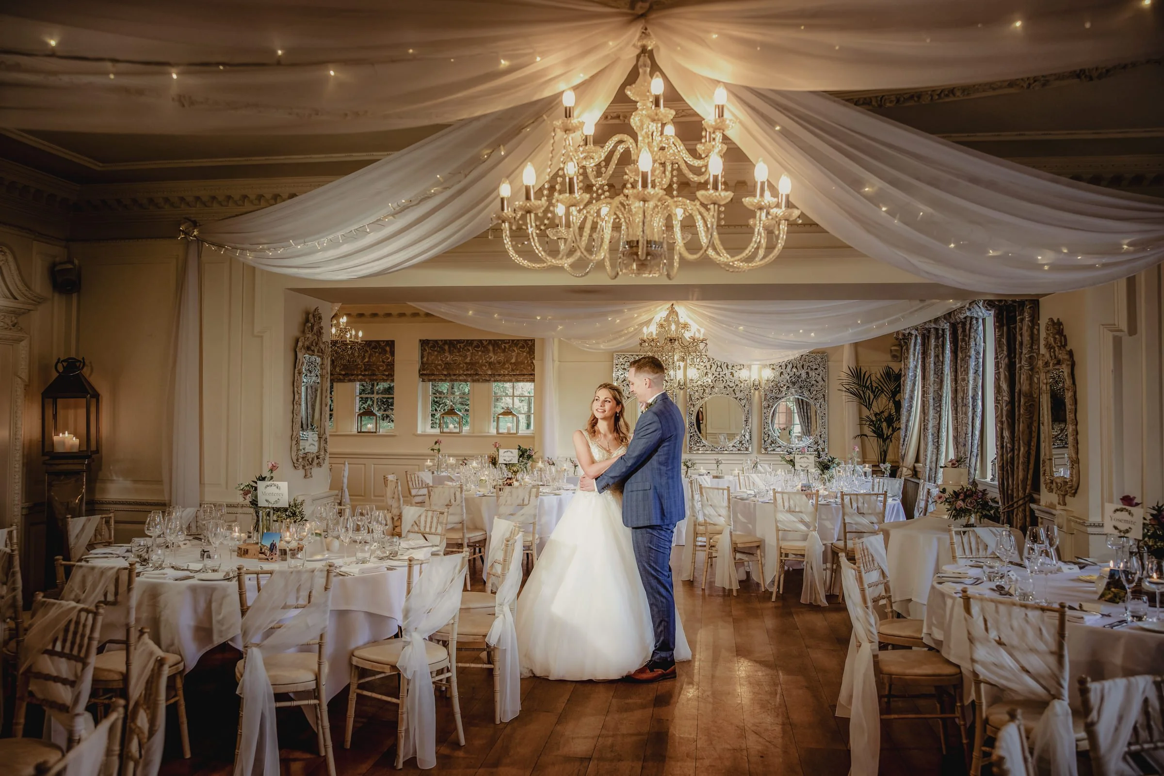 Bride and groom dancing in a decorated wedding reception hall with chandeliers, white drapes, and round tables set for a celebration.