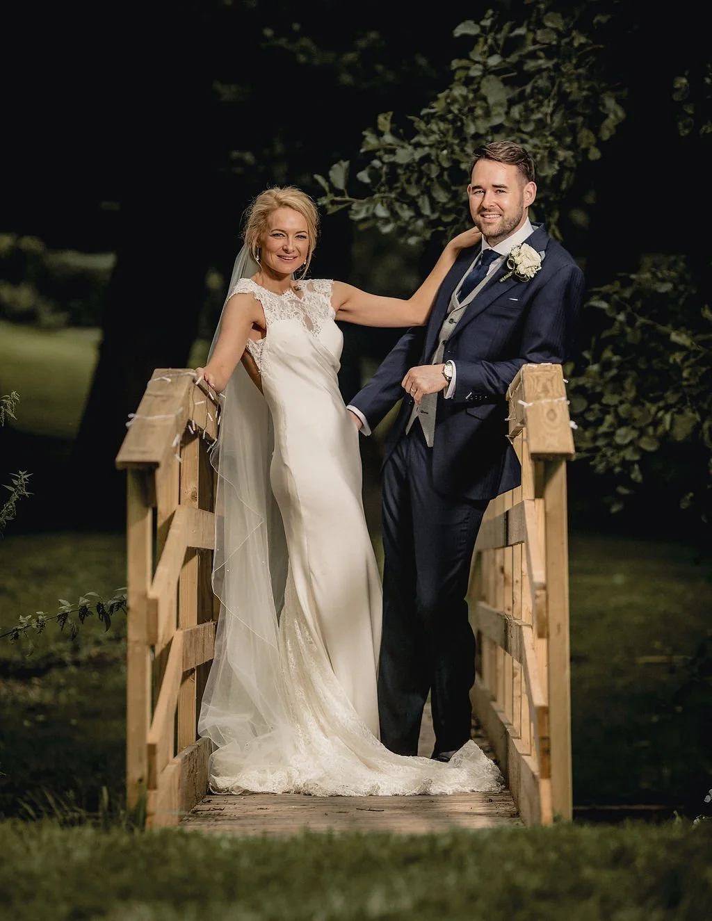 Wedding couple standing on a small wooden bridge outdoors, smiling at the camera, with trees and greenery in the background.