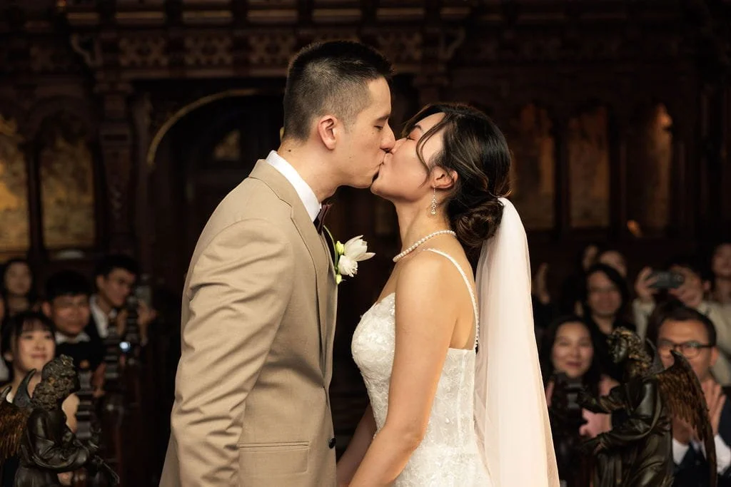 A bride and groom kiss during their wedding ceremony as guests look on and take photos.