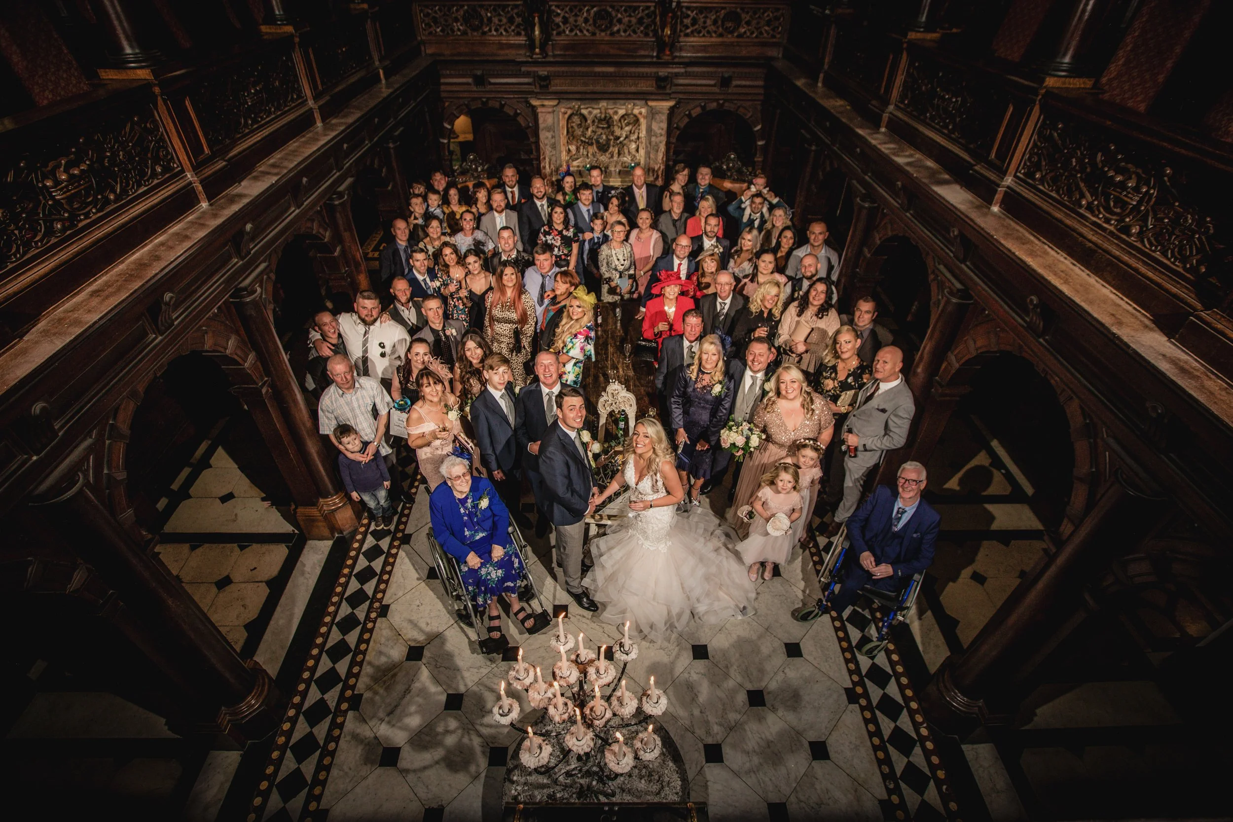 A wedding celebration with a bride and groom holding hands in the center, surrounded by guests in a grand, ornate hall with dark wood accents and a large chandelier.