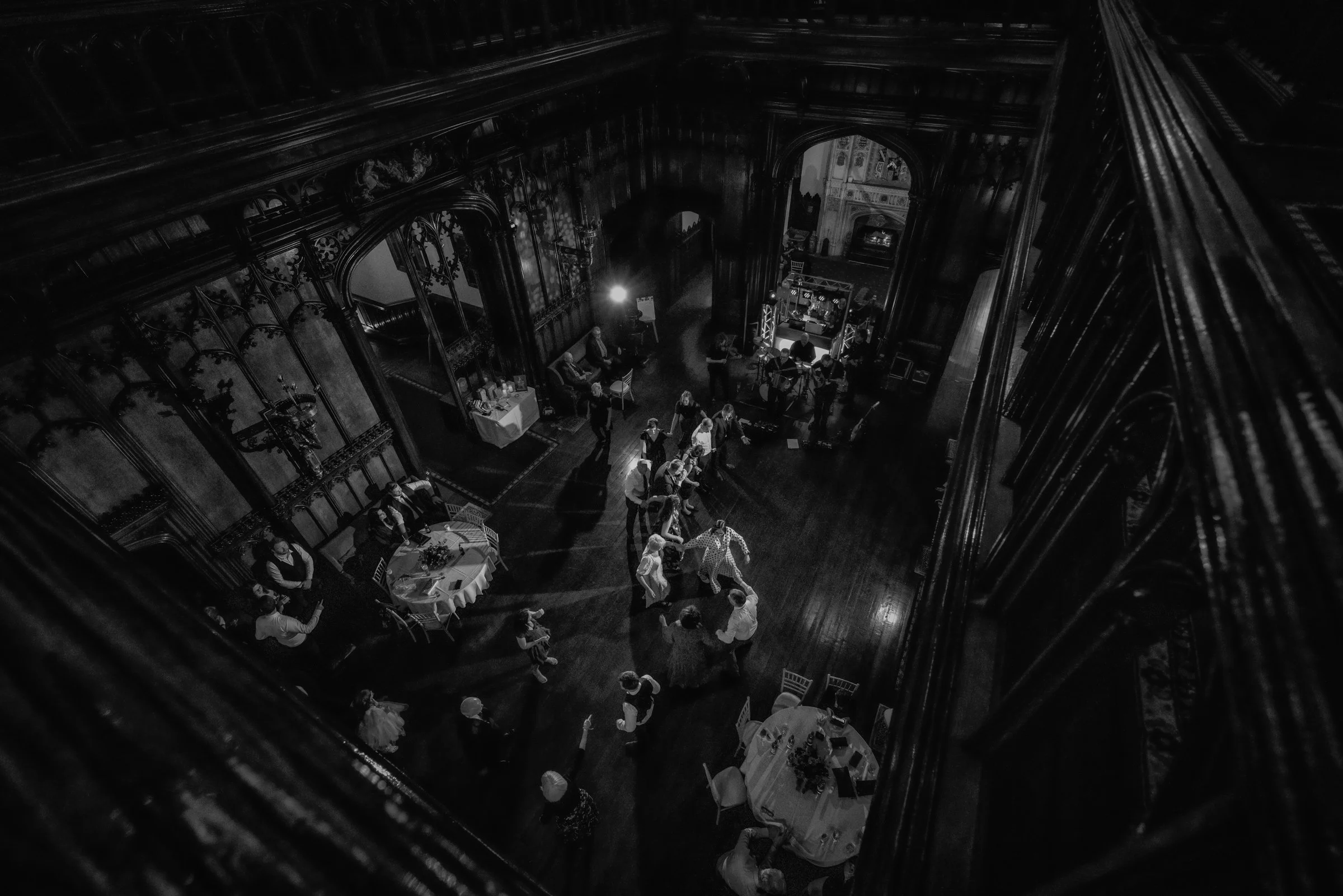 People dancing and socializing at an event in an elegant, wood-paneled hall with high ceilings and ornate architectural details, viewed from an elevated angle.