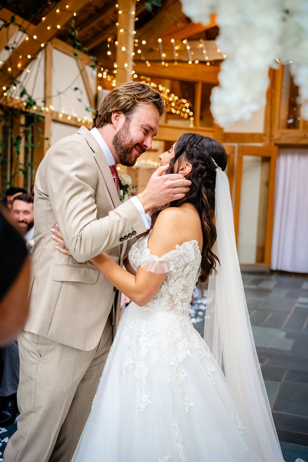 A bride and groom share a joyful moment during their wedding ceremony. The groom is gently holding the bride's face as they smile at each other in a decorated indoor venue with warm lighting.