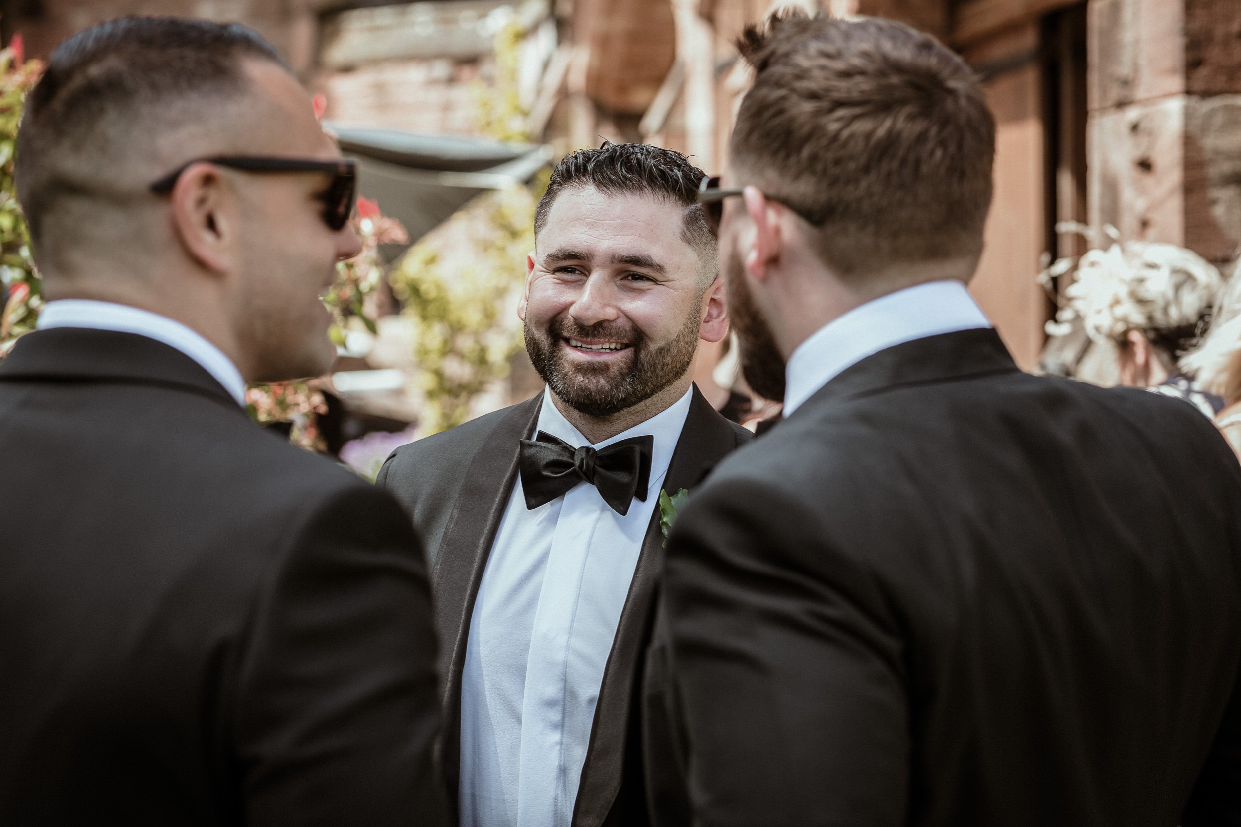 Three men in tuxedos and bow ties standing together at an outdoor wedding, smiling and having a conversation.