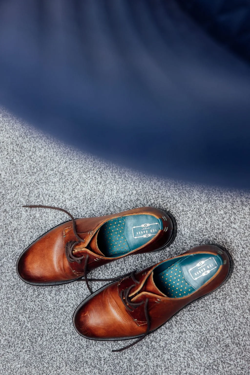 Brown leather dress shoes with blue insoles on a gray carpeted floor next to a navy blue fabric.