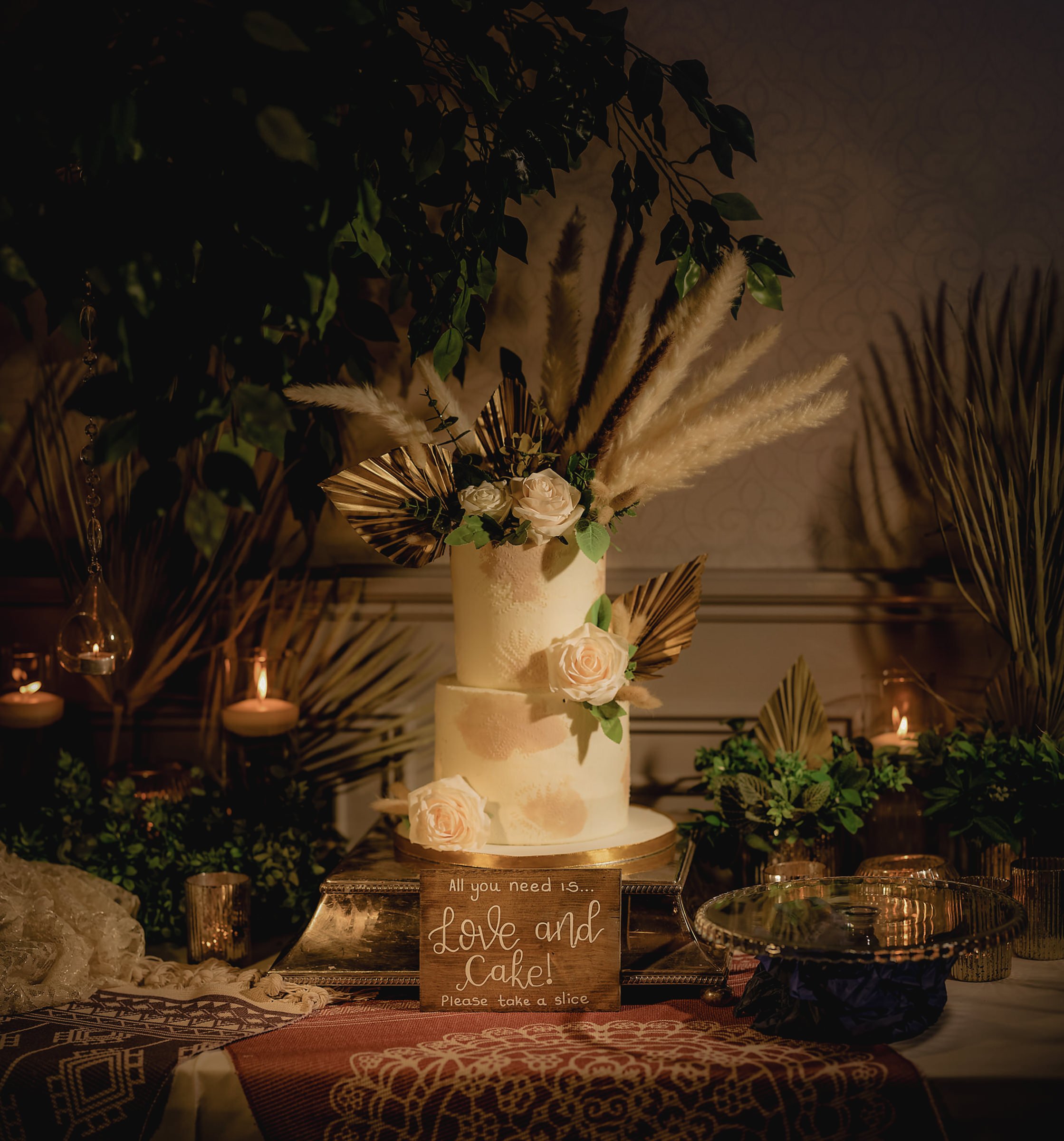 Decorative wedding cake with flowers and foliage, displayed on a table with candles and greenery in the background.
