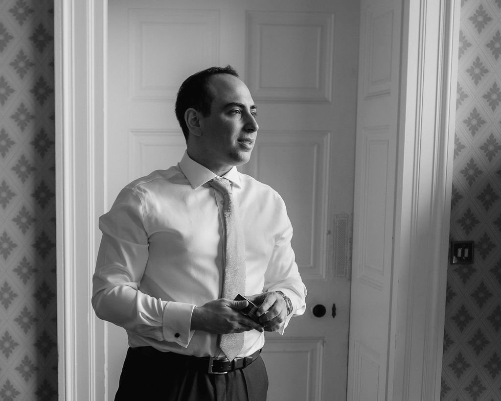 A man in a white shirt and tie standing in front of a door, holding a phone, in black and white.