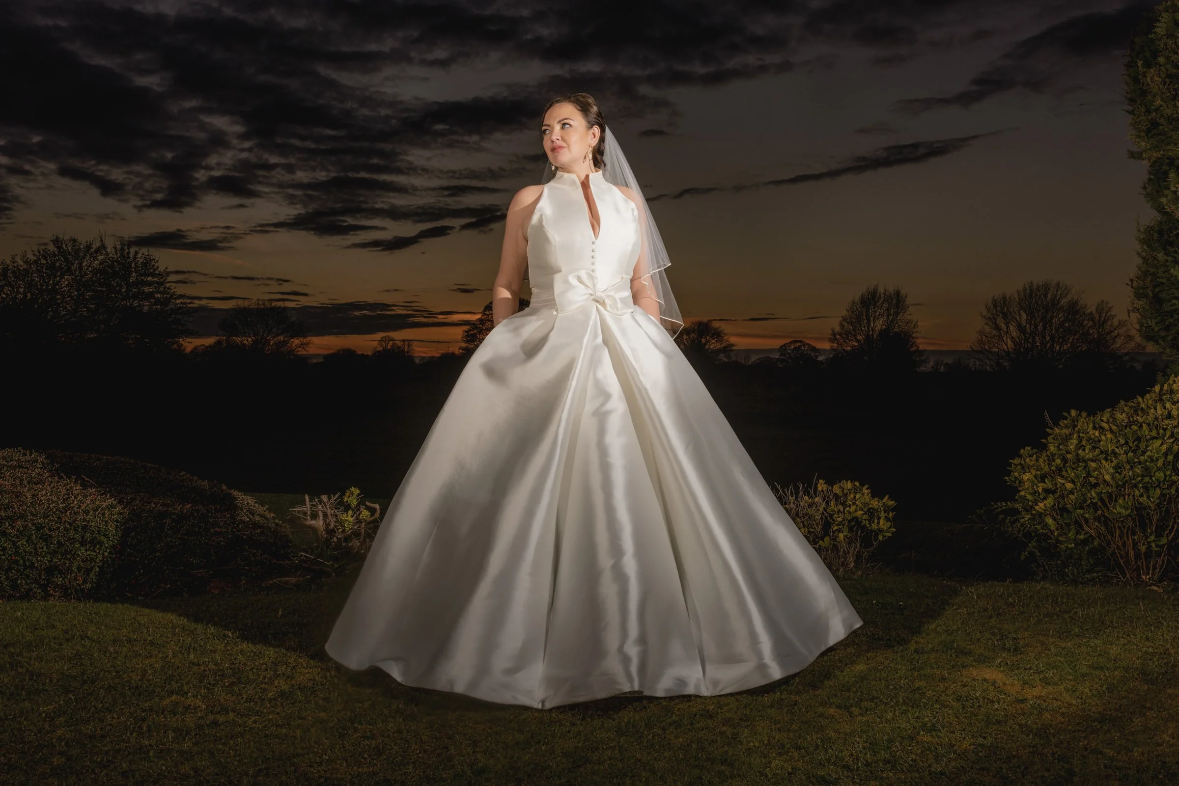 A woman in a white wedding gown with a veil standing outdoors at sunset with an orange and dark sky, surrounded by greenery and trees.