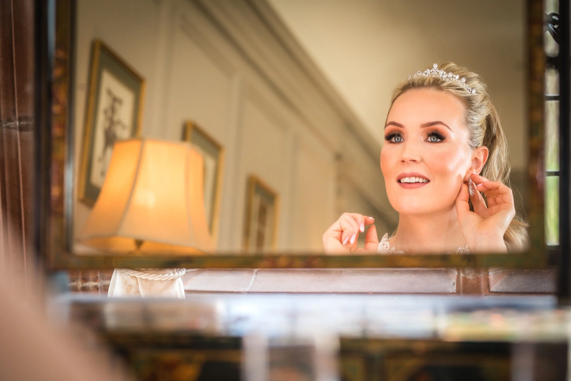Bride in a white wedding dress, with a tiara, putting on an earring while looking in a mirror in a warmly lit room.