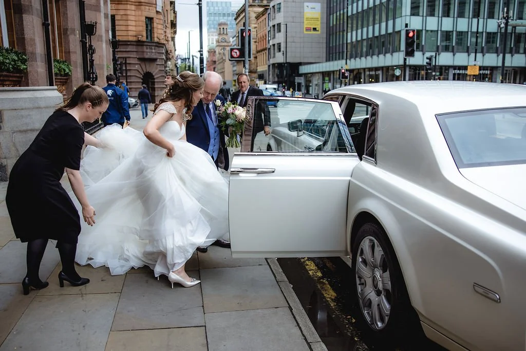 A bride in a white wedding dress getting into a luxury car with the help of an older man, while a woman in black assists with her dress on a city sidewalk.