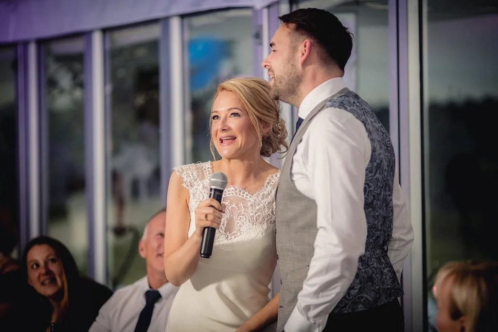 A woman giving a speech at a wedding reception while holding a microphone, standing next to a man in formal attire, with wedding guests smiling in the background.