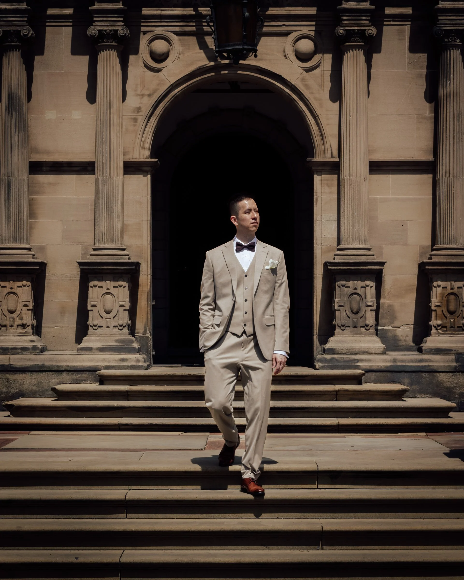 A man in a beige suit with a bow tie and white shirt, standing on steps in front of an ornate stone building with columns and an arched entrance.