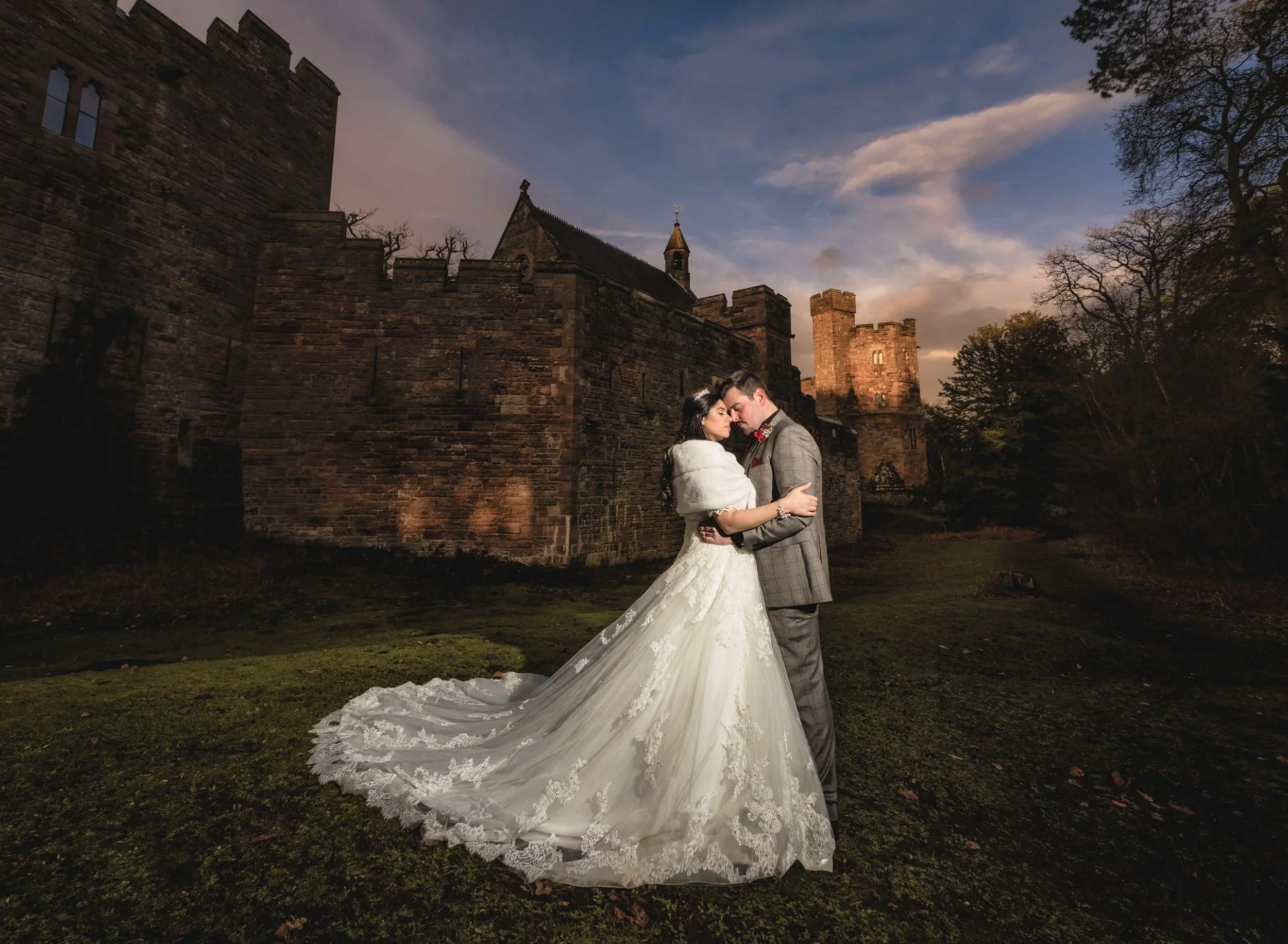 A bride and groom embracing outdoors at a sunset, with a castle and trees in the background.