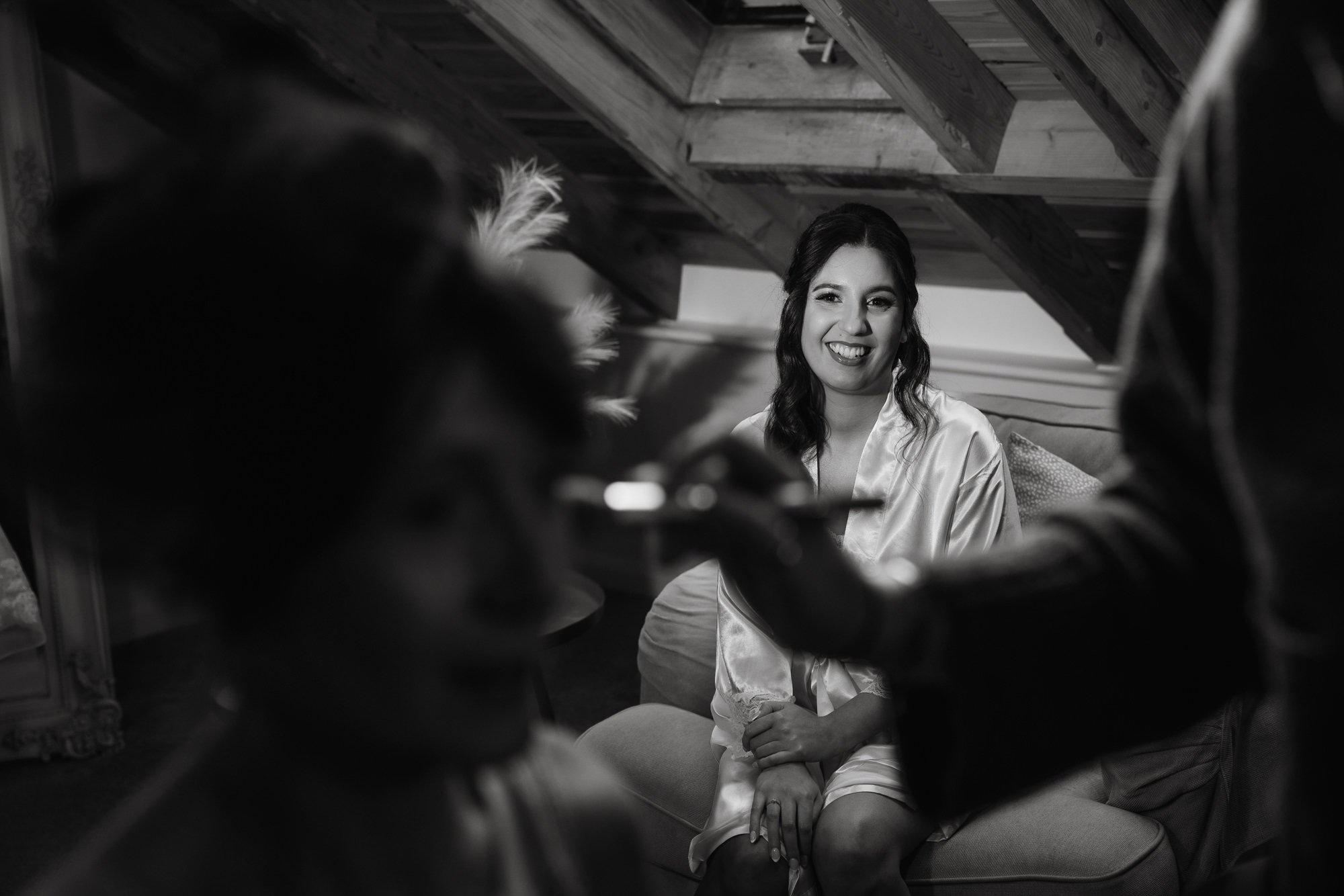 A woman smiling in a satin robe having makeup applied by a makeup artist, with a person blurred in the foreground, in a cozy attic space with wooden beams.