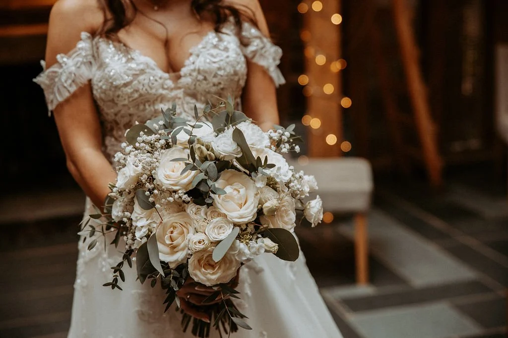 Bride in a lace wedding dress holding a bouquet of white roses, baby's breath, and greenery in a rustic indoor setting with string lights in the background.