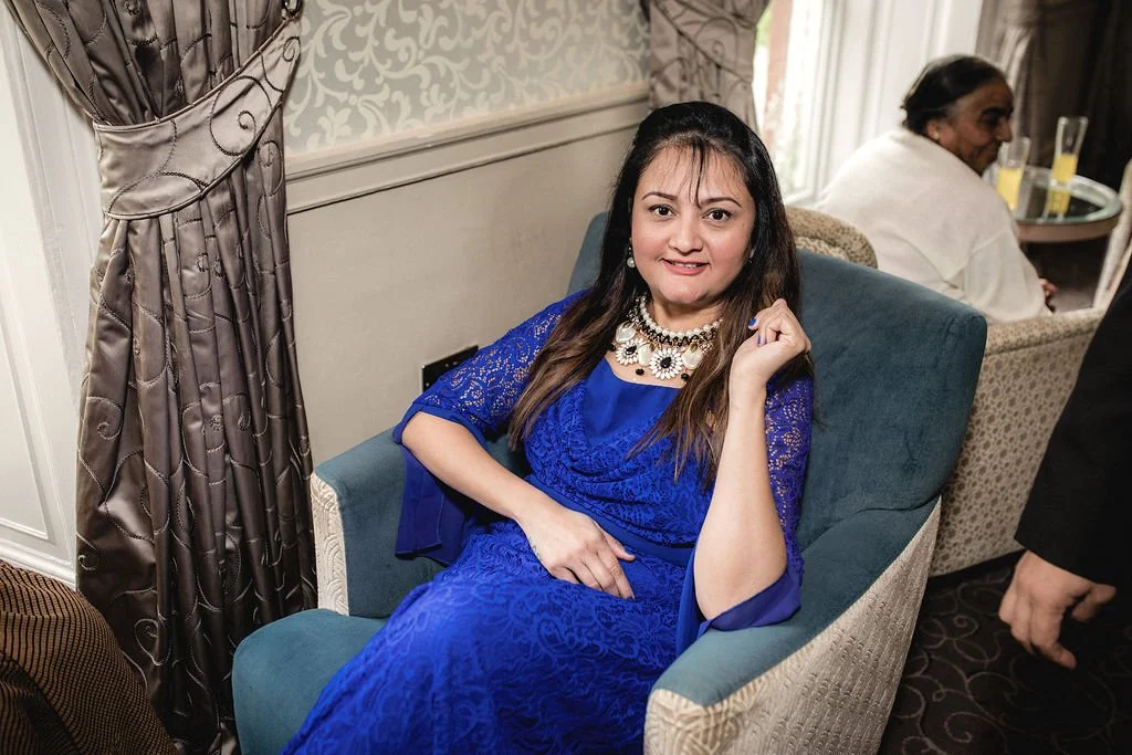 A woman in a royal blue dress and necklace sitting on a chair at a social gathering, with a woman in a white outfit seated in the background.