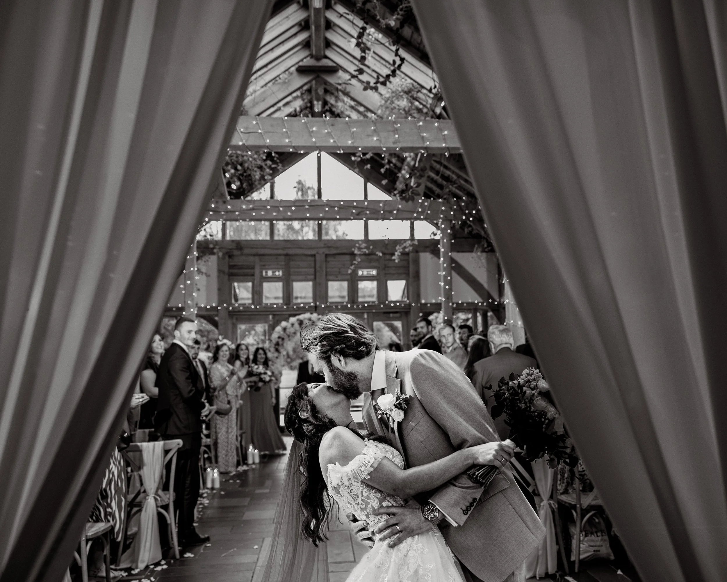 A couple sharing a kiss during their wedding ceremony inside a decorated venue, with guests watching in the background.