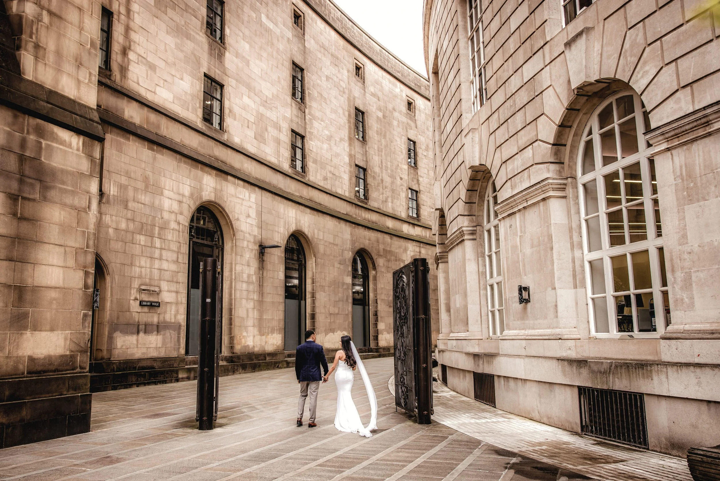 A bride and groom holding hands, walking through an open gate in a historic stone building courtyard.