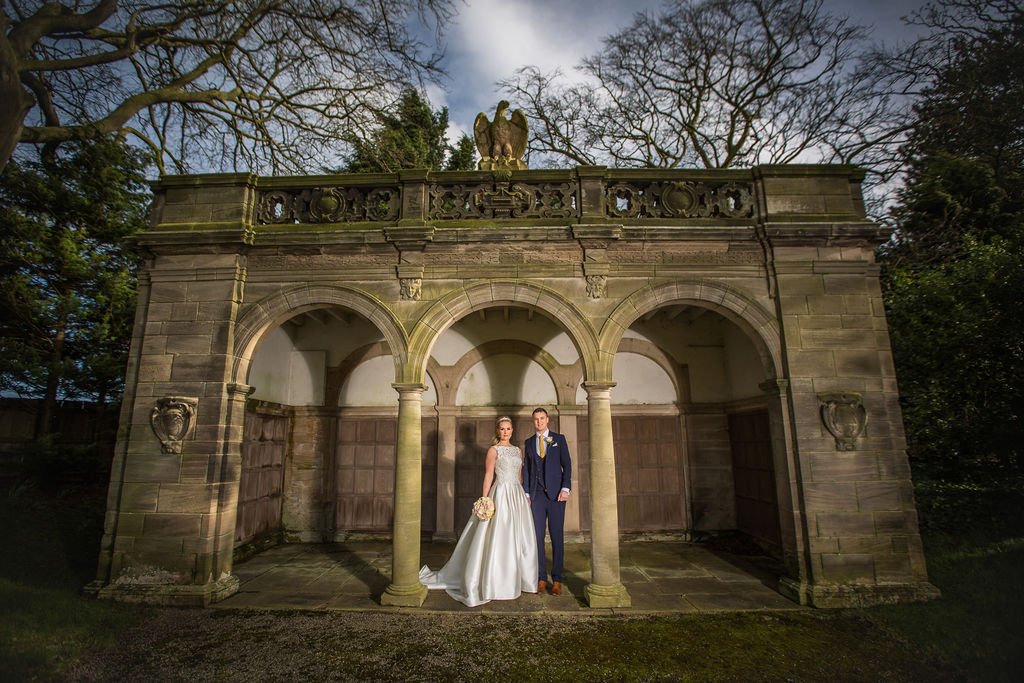 A bride and groom standing together beneath a stone archway with a decorative railing and an eagle statue on top, outdoors surrounded by trees.