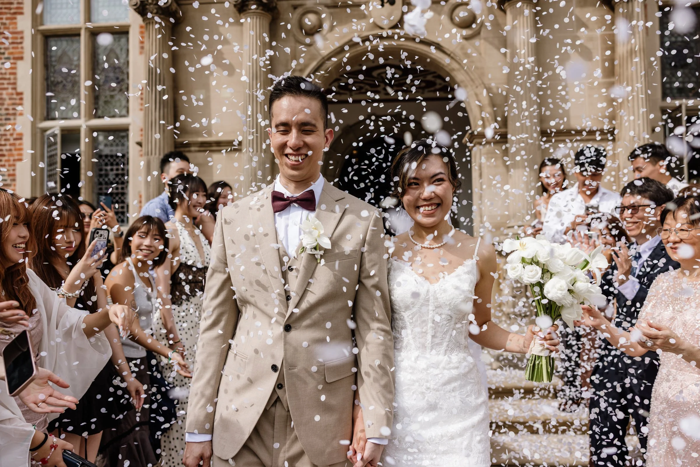 A newly married couple smiling and holding hands after their wedding ceremony, surrounded by friends and family throwing white confetti outside a historic building.