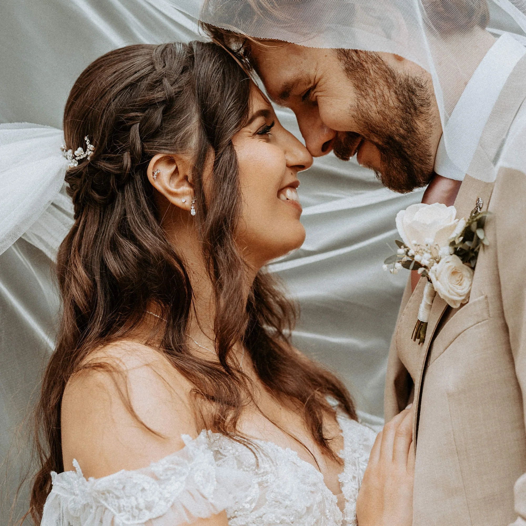 A bride and groom softly touching foreheads and closing their eyes during a wedding ceremony, with the bride wearing a lace wedding dress and the groom in a tan suit holding a bouquet of white roses.