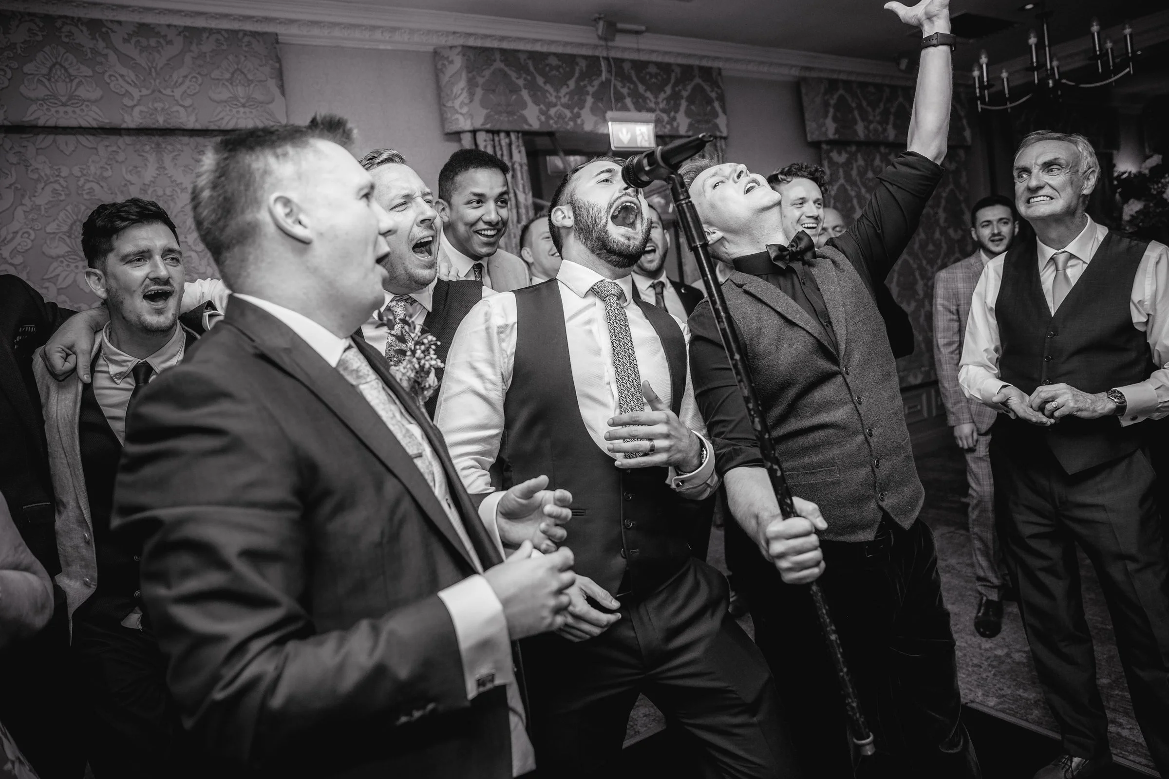 Group of men singing and dancing at an indoor celebration, some with smiles and animated expressions, microphone stand present, formal attire