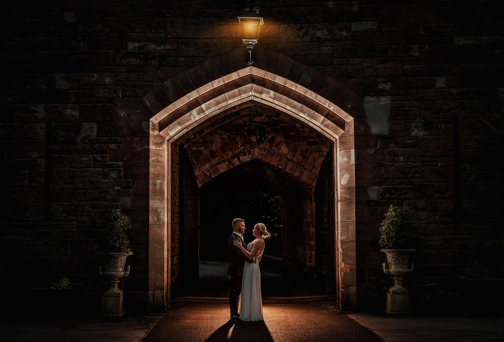 A bride and groom stand close together under a lit archway at night, holding hands and looking into each other's eyes.