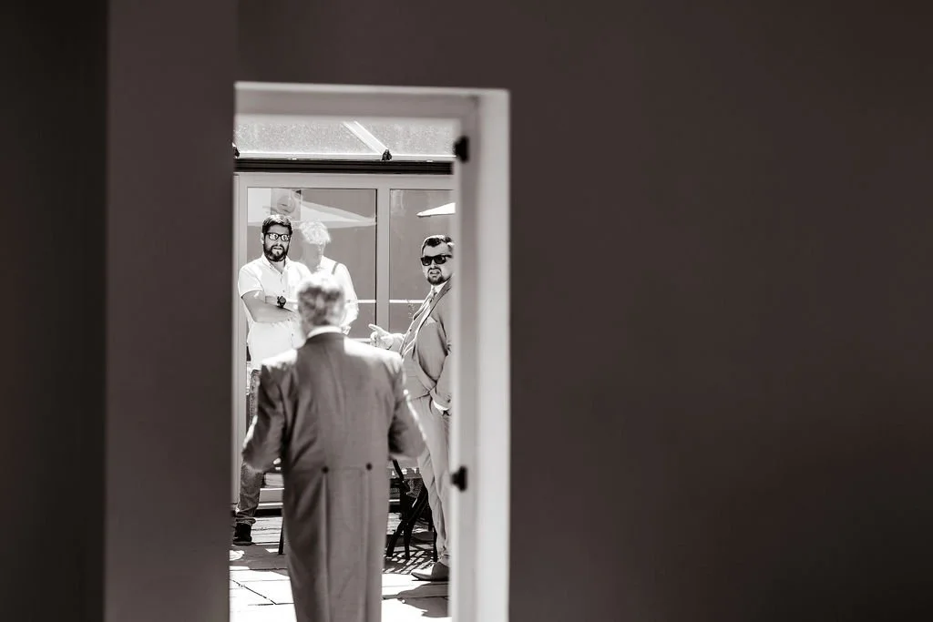 Group of four men standing outside, seen through a doorway, with one man in the foreground facing away and three men facing towards the camera, two of them wearing sunglasses.