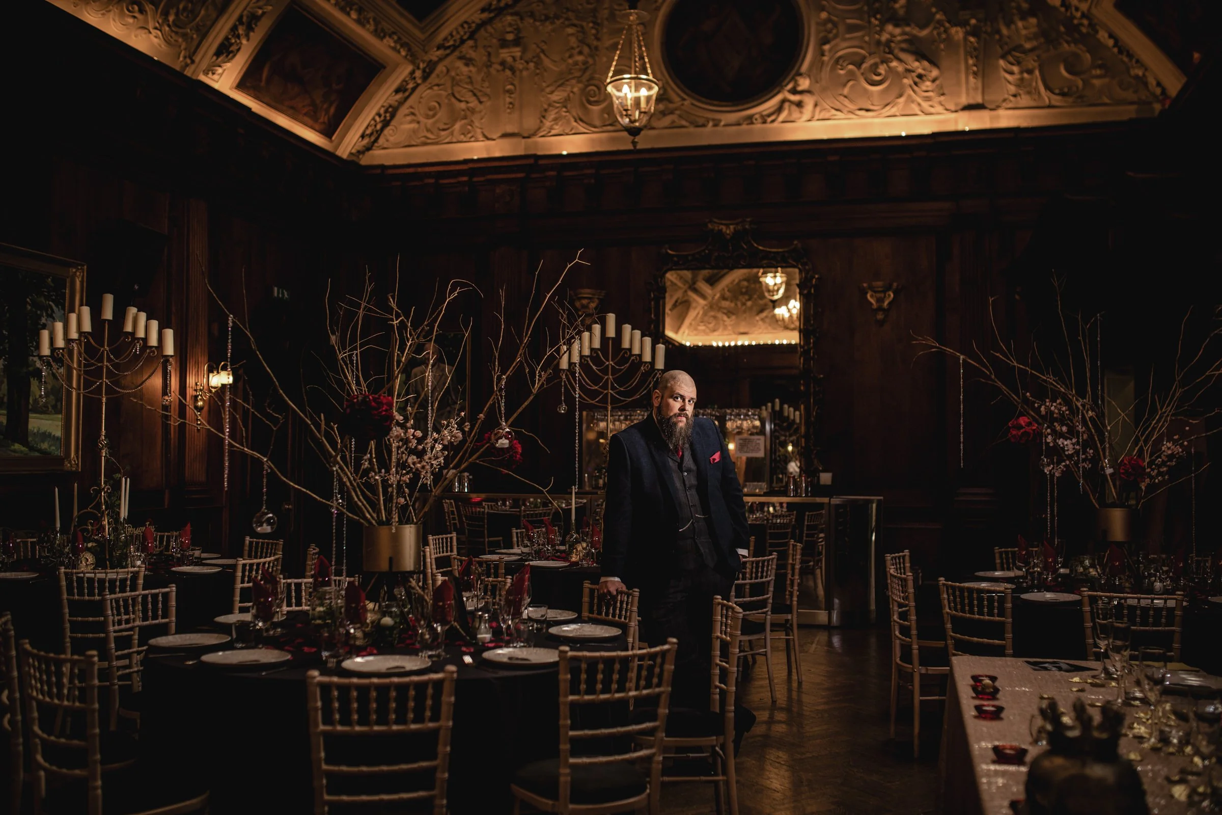 A man with a beard dressed in a dark suit stands in a dimly lit, ornate event hall with dark wood paneling. Elegant tables with black tablecloths are set with glassware and red napkins, and large floral arrangements with bare branches and red flowers