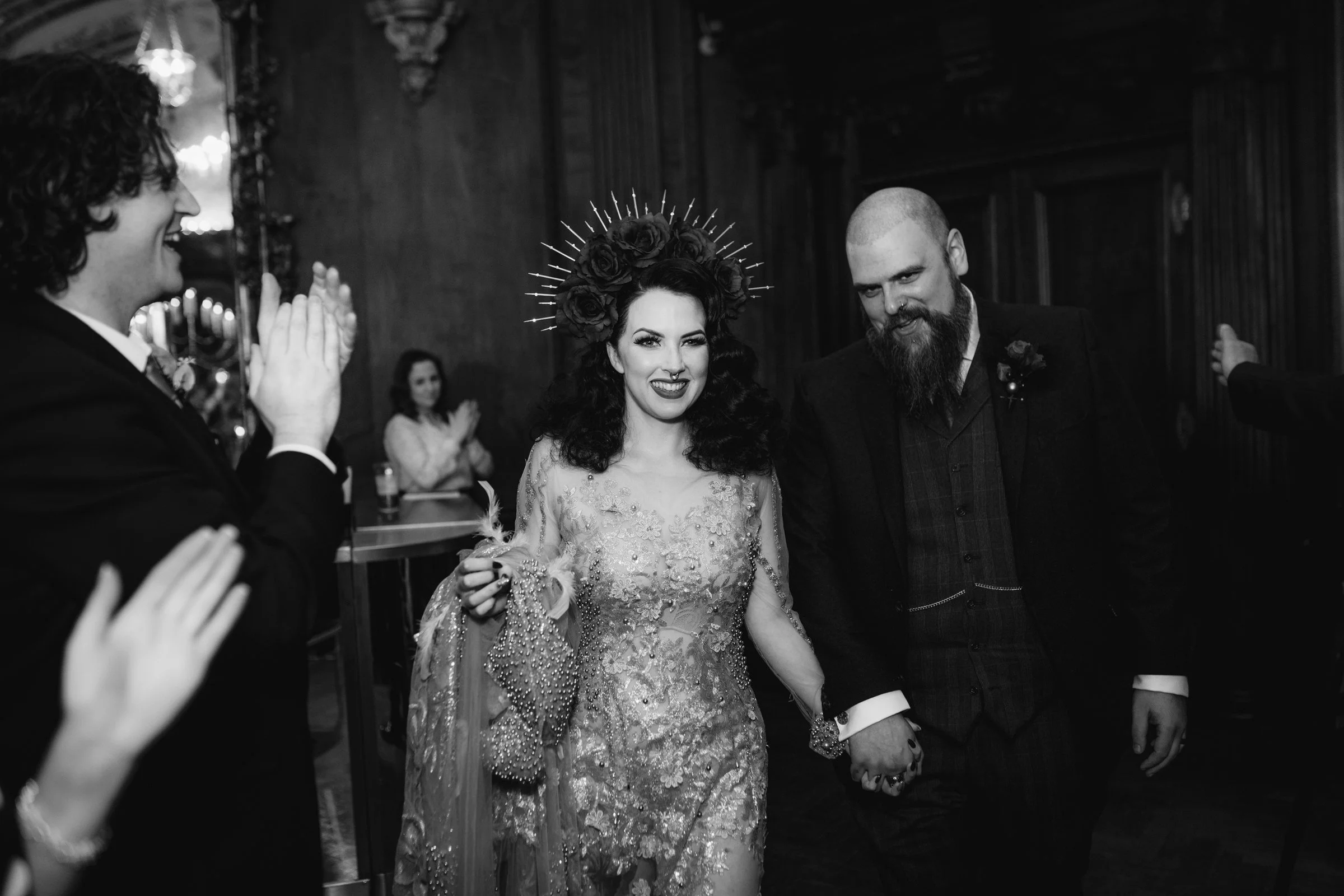 Black and white photo of a wedding reception featuring a bride wearing an ornate dress and a floral headpiece, holding the hand of a bearded groom in a suit. Guests are clapping and smiling in the background.