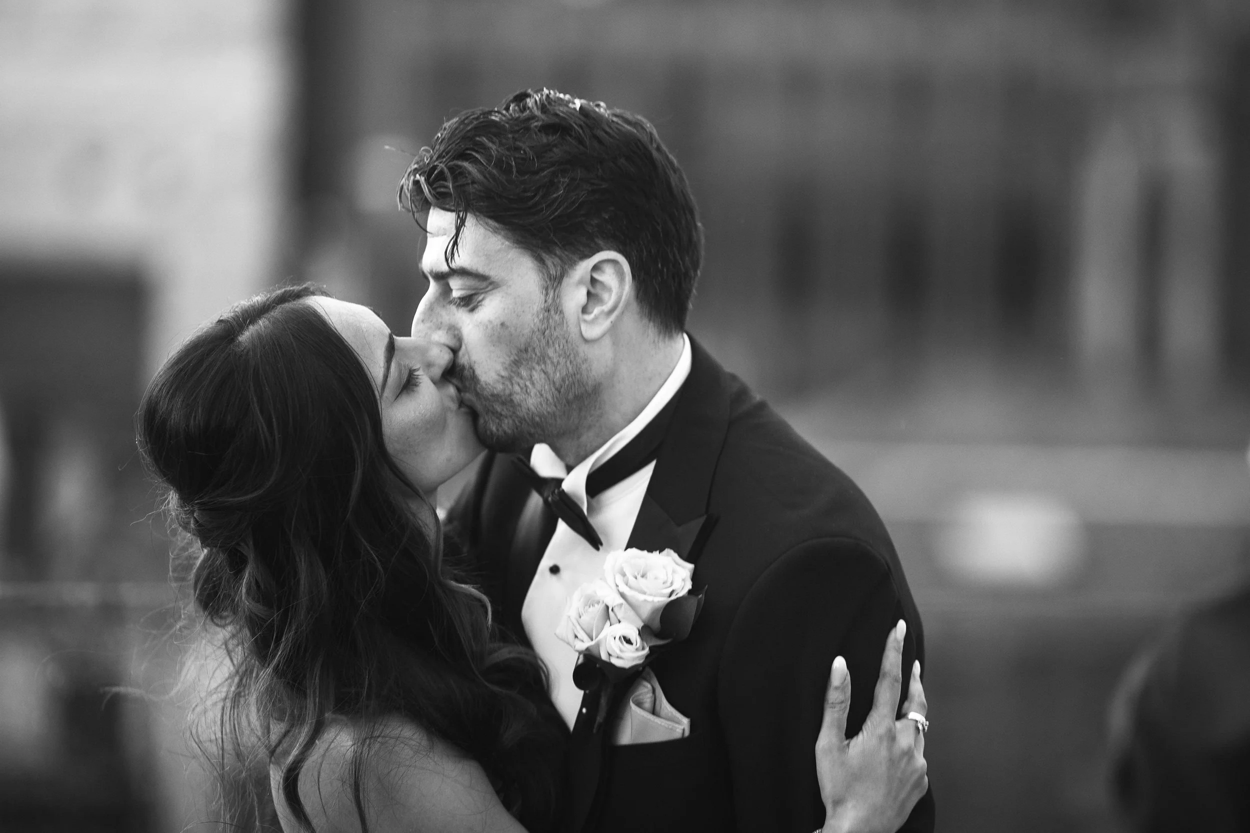 A black and white photo of a couple kissing, dressed in formal wedding attire, with the groom wearing a tuxedo and boutonniere.