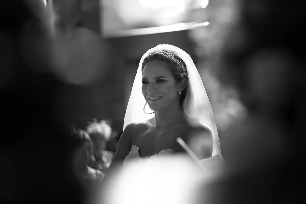 A black and white photograph of a smiling bride with a veil and tiara, looking serene in a wedding dress, surrounded by blurred figures.