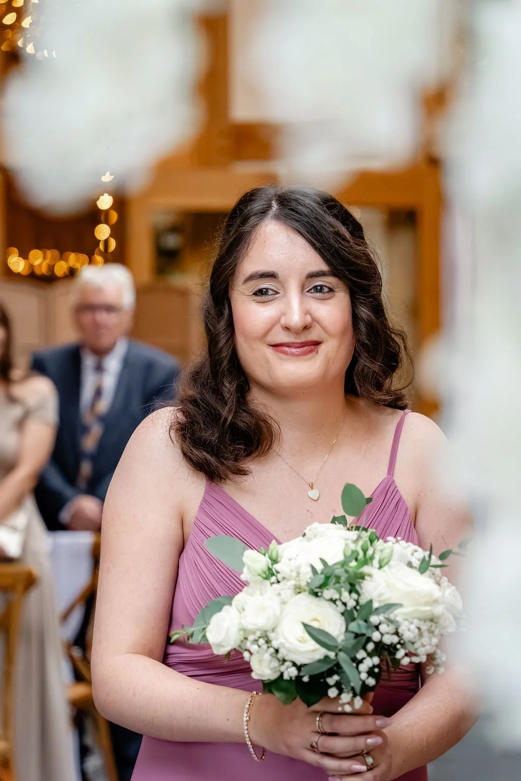 A woman in a pink dress holding a bouquet of white flowers at a wedding ceremony. She is smiling and wearing jewelry, with blurred guests in the background.