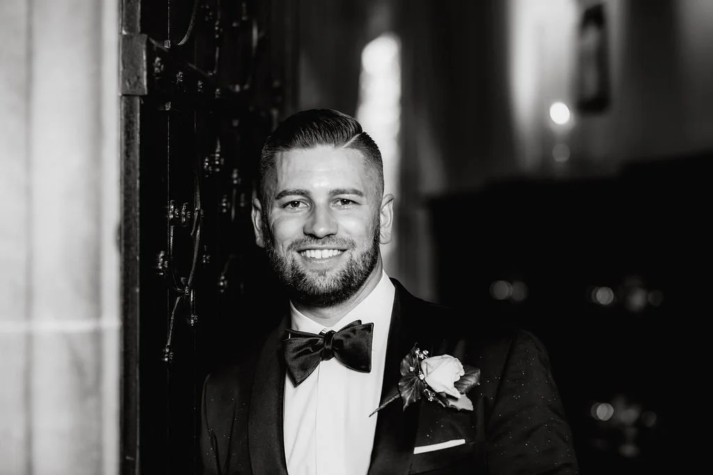 A man in a tuxedo with a bow tie and boutonniere, smiling in a black and white photo.