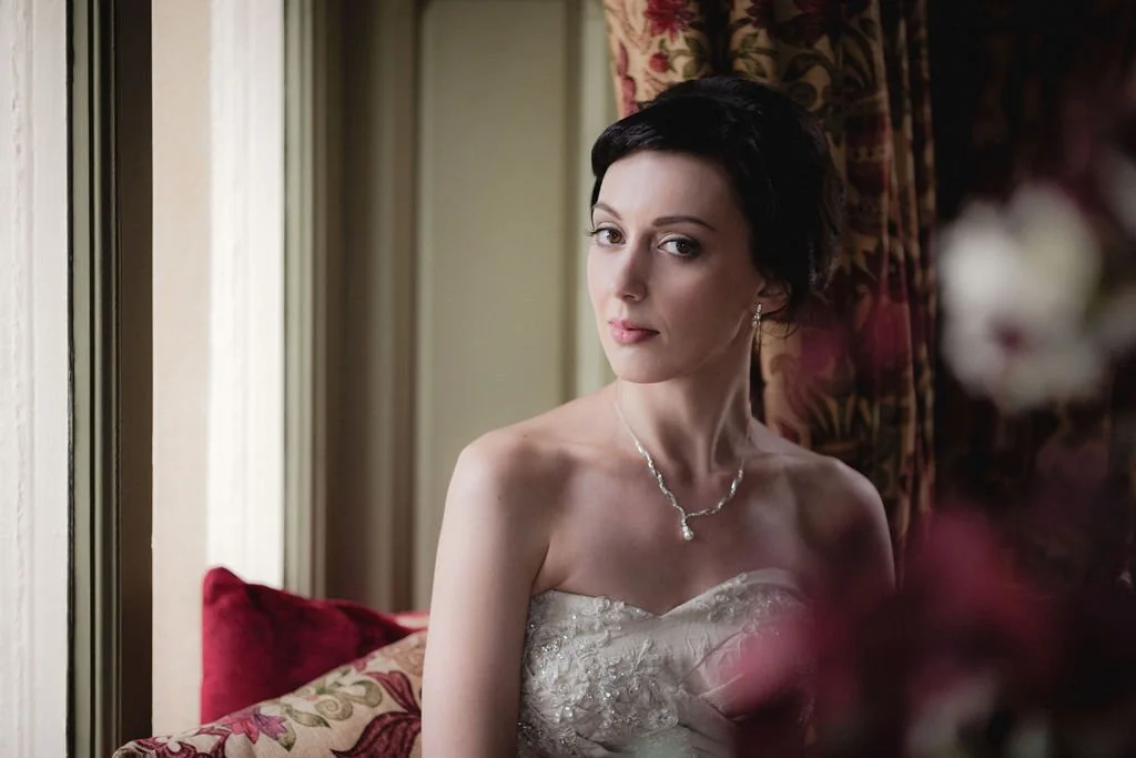 A woman with short dark hair and fair skin wearing a strapless lace dress and jewelry, sitting by a window with curtains, looking at the camera.