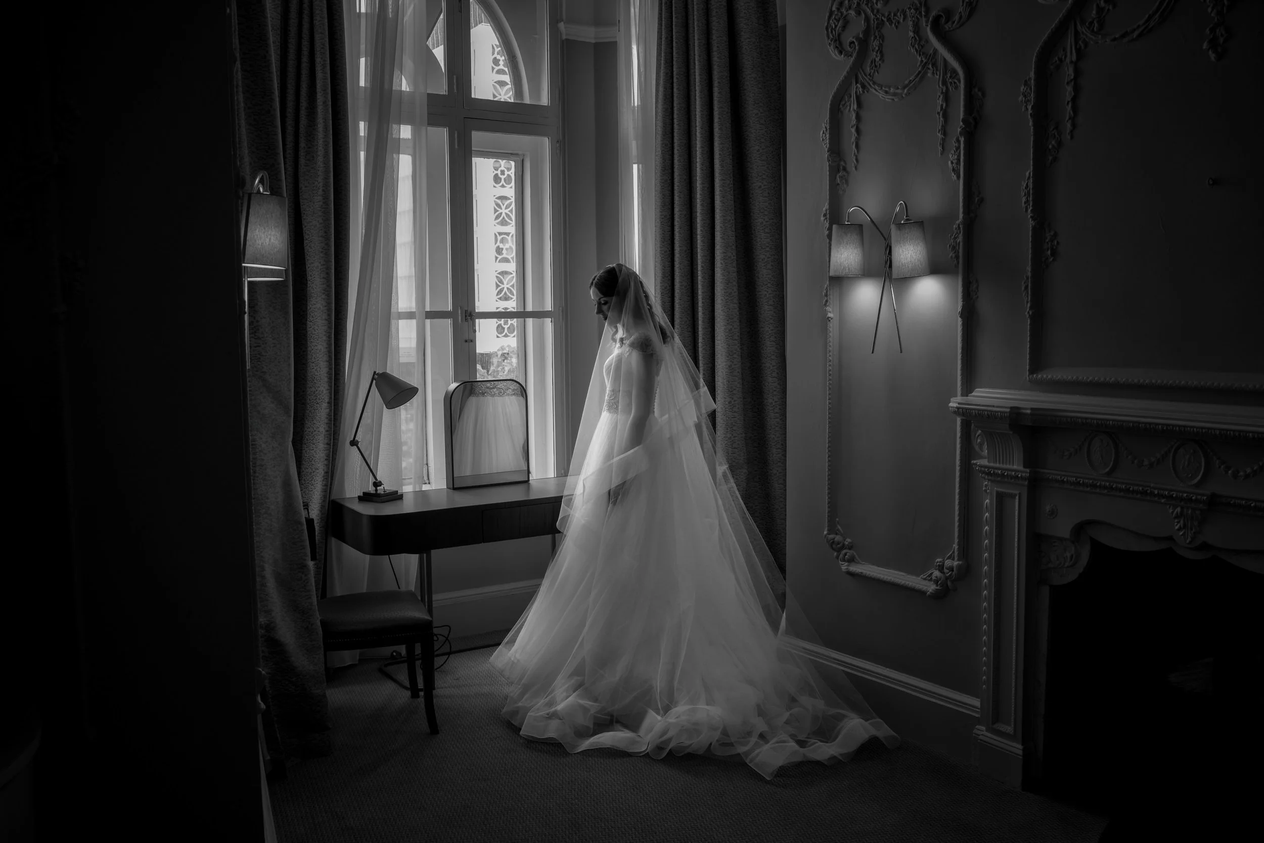A bride in a wedding dress and veil standing by a window in a dimly lit room with elegant decor, contemplating.
