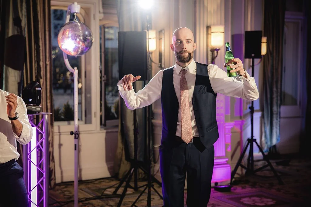 A man with a bald head and beard wearing a suit with a pink tie, holding a beer bottle in one hand and dancing at a party with disco lights and decor.