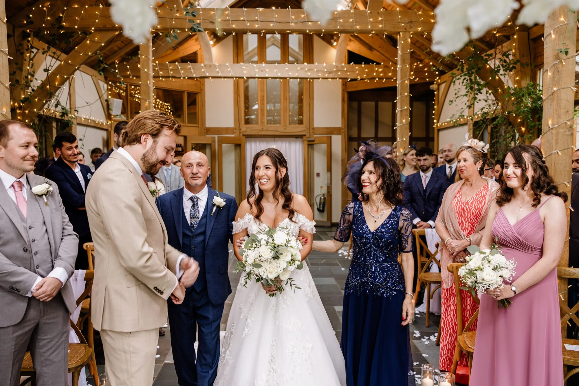 A wedding ceremony with the bride and groom smiling, surrounded by family and friends in a decorated indoor venue with string lights and wooden beams.