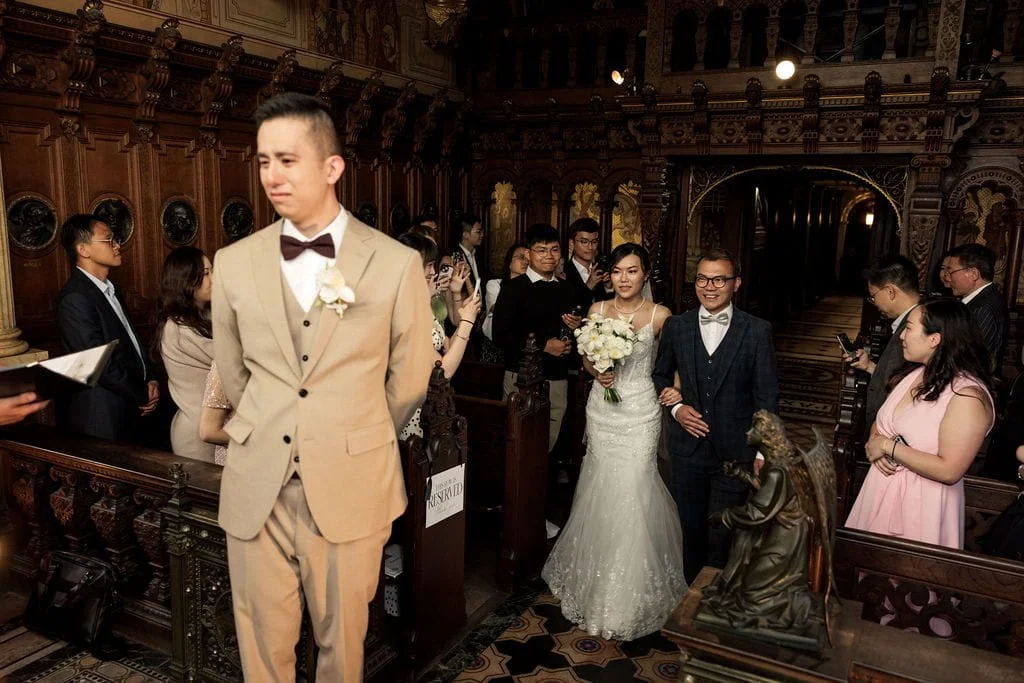 Bride and groom walking down the aisle in a church with guests on both sides, bride holding a bouquet of white flowers, groom wearing glasses and a dark suit, wedding party and guests in formal attire, ornate wooden church interior.