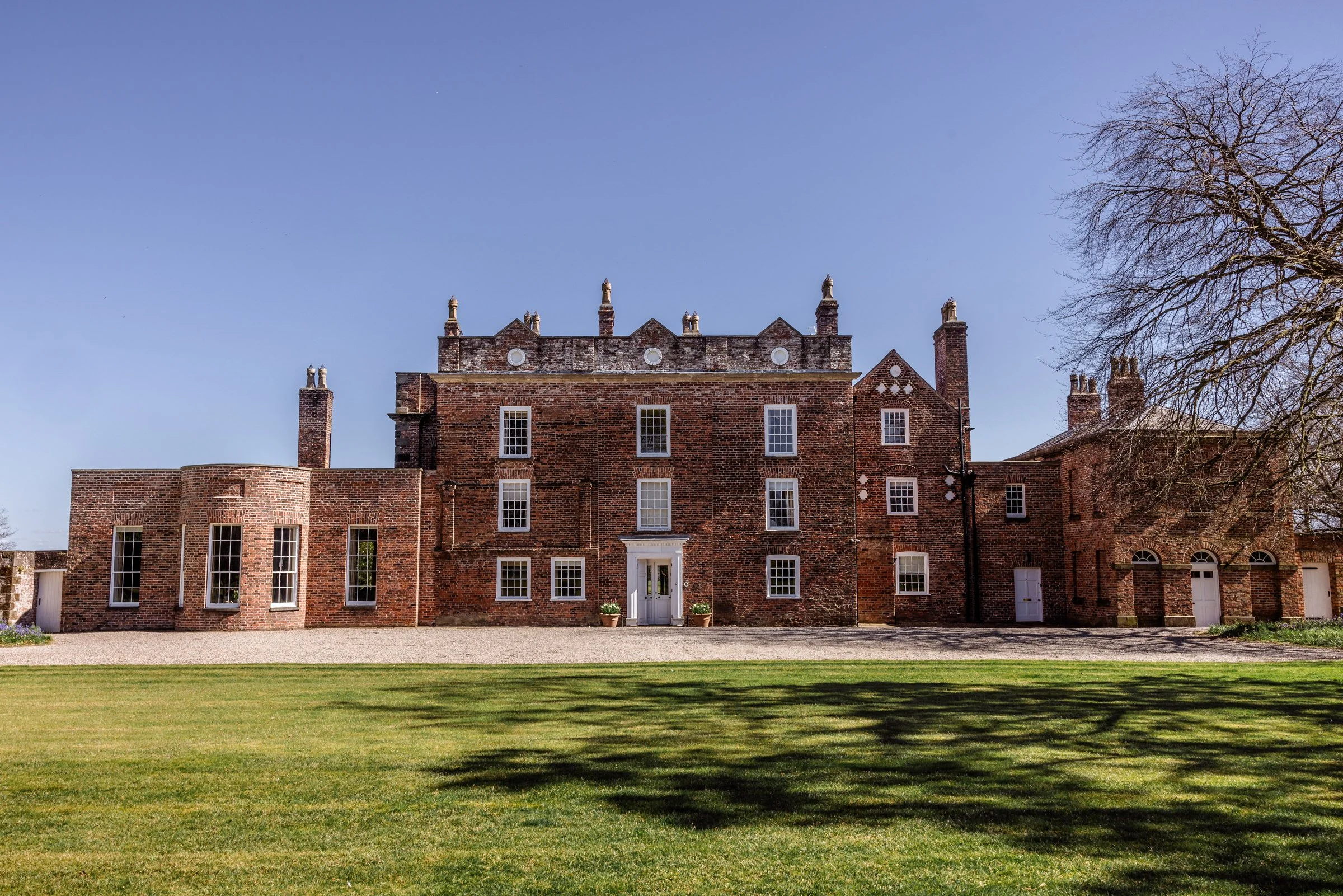 A large, historic brick mansion with multiple stories, white-framed windows, and chimneys, surrounded by a well-maintained lawn and trees under a clear blue sky.